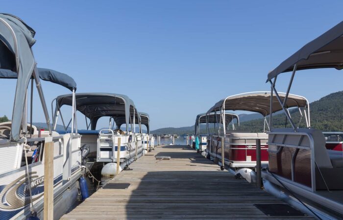 A wooden dock lined with pontoon boats on both sides, under a clear blue sky, with forested hills visible in the background.