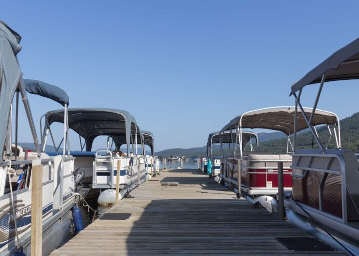 A wooden dock lined with pontoon boats on both sides, under a clear blue sky, with forested hills visible in the background.