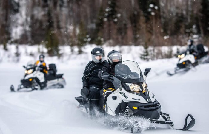 Four people ride snowmobiles through a snowy forest, wearing helmets and winter gear, with snow and trees in the background.