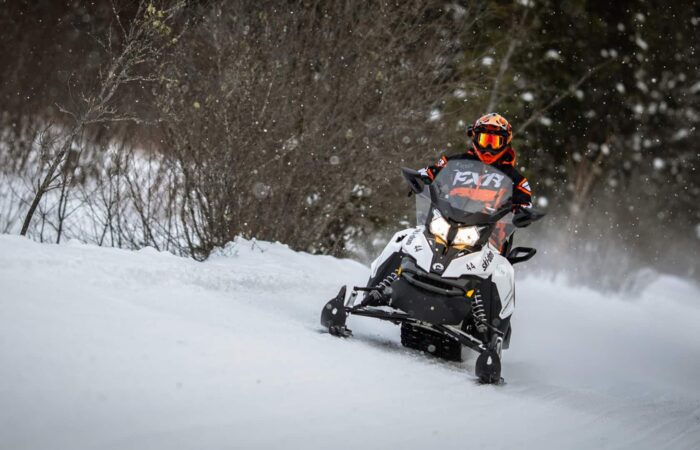 Personne conduisant une motoneige sur un sentier enneigé entouré d'arbres, portant un casque et un équipement d'hiver - parfait pour les amateurs d'aventure recherchant des sensations fortes lors de leurs activités Mont-Tremblant.