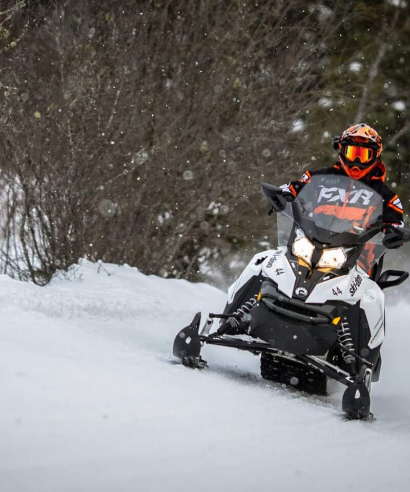 Personne conduisant une motoneige sur un sentier enneigé entouré d'arbres, portant un casque et un équipement d'hiver - parfait pour les amateurs d'aventure recherchant des sensations fortes lors de leurs activités Mont-Tremblant.