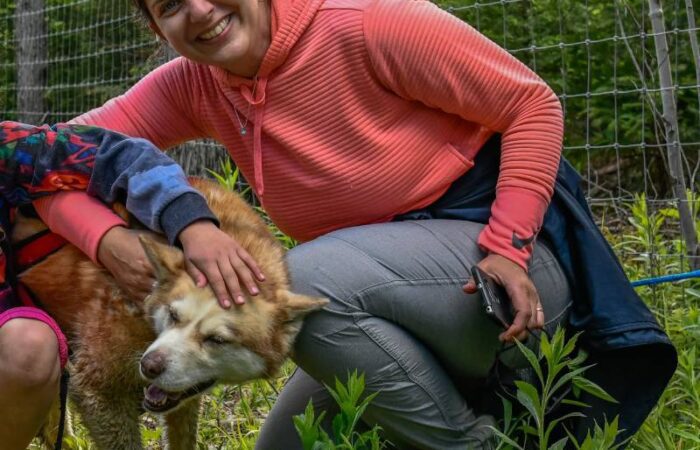 A woman in a pink hoodie smiles while crouching next to a tan and white dog, petting its head in a grassy outdoor area with a wire fence in the background.