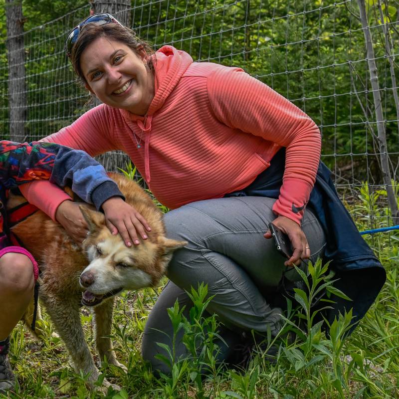 A woman in a pink hoodie smiles while crouching next to a tan and white dog, petting its head in a grassy outdoor area with a wire fence in the background. A woman in a pink hoodie smiles while crouching next to a tan and white dog, petting its head in a grassy outdoor area with a wire fence in the background.