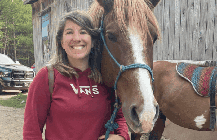 A woman in a red Vans hoodie stands smiling next to a brown horse with a blue halter, in front of a wooden building.