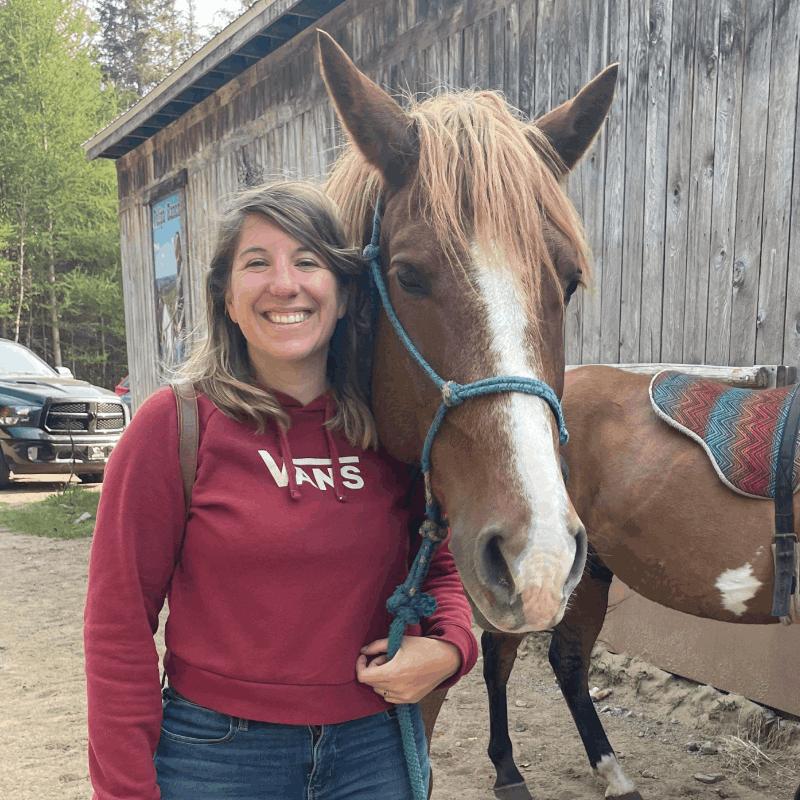 A woman in a red Vans hoodie stands smiling next to a brown horse with a blue halter, in front of a wooden building. A woman in a red Vans hoodie stands smiling next to a brown horse with a blue halter, in front of a wooden building.