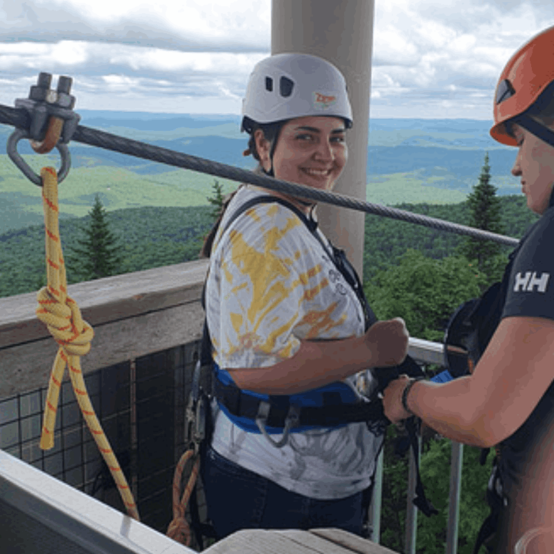 A person wearing a helmet and harness prepares for a zipline ride with the help of an instructor on a platform overlooking forested mountains. A person wearing a helmet and harness prepares for a zipline ride with the help of an instructor on a platform overlooking forested mountains.