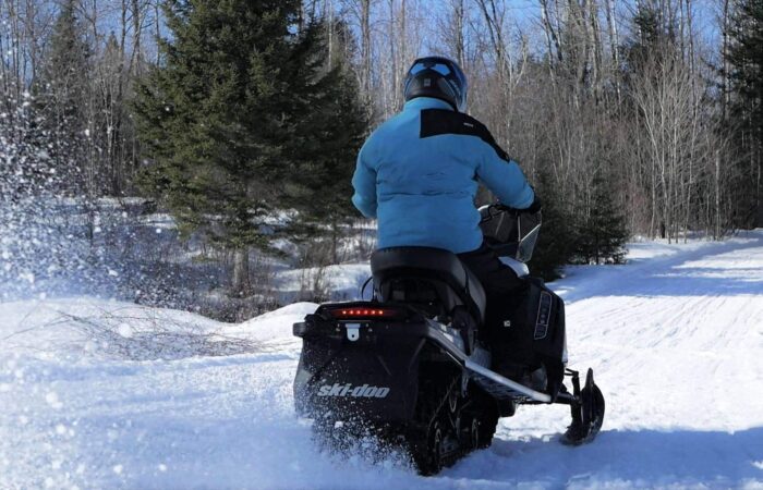 A person wearing a blue jacket and helmet rides a black snowmobile on a snowy trail surrounded by leafless trees.