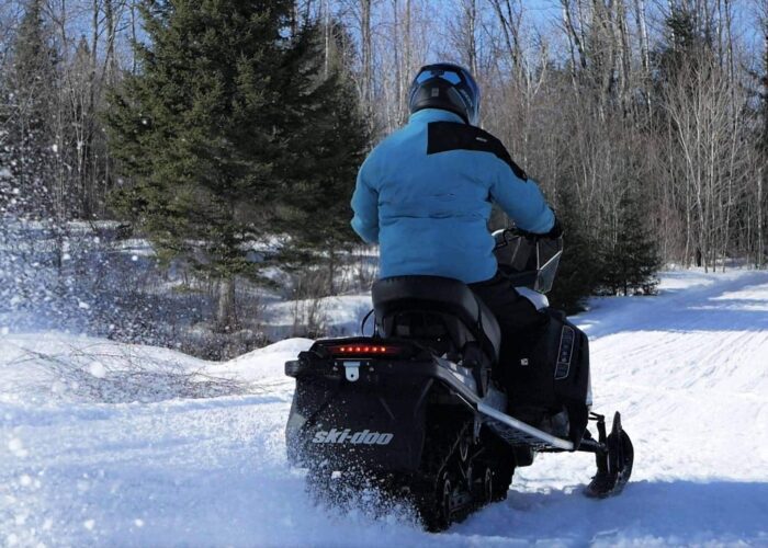 A person wearing a blue jacket and helmet rides a black snowmobile on a snowy trail surrounded by leafless trees.