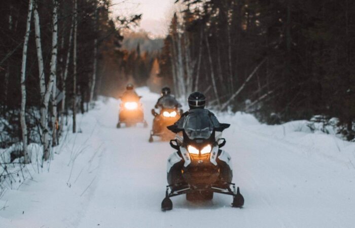 Three people ride snowmobiles on a snowy forest trail, with headlights on and trees lining both sides of the path.
