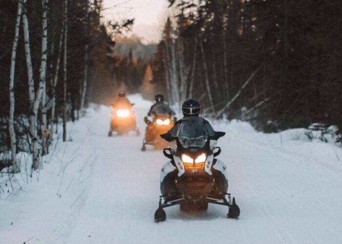 Three people ride snowmobiles on a snowy forest trail, with headlights on and trees lining both sides of the path.
