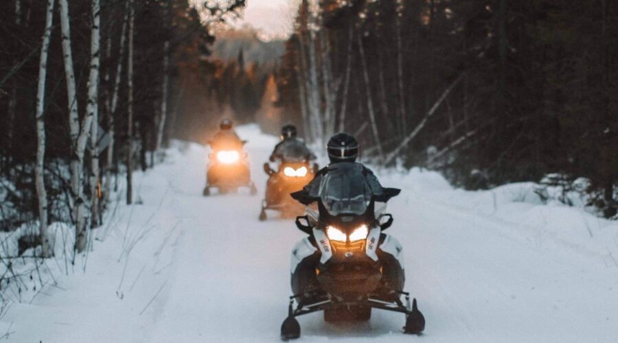Three people ride snowmobiles on a snowy forest trail, with headlights on and trees lining both sides of the path. Three people ride snowmobiles on a snowy forest trail, with headlights on and trees lining both sides of the path.