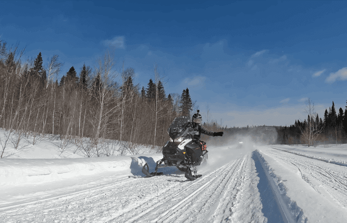 A person rides a snowmobile on a snowy trail through a forested area under a clear blue sky.
