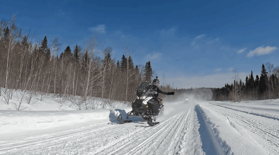 A person rides a snowmobile on a snowy trail through a forested area under a clear blue sky. A person rides a snowmobile on a snowy trail through a forested area under a clear blue sky.