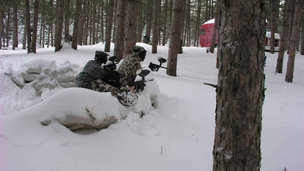 Three people in camouflage gear crouch behind a snow-covered barrier in a snowy forest, holding paintball markers, with a red structure visible in the background.