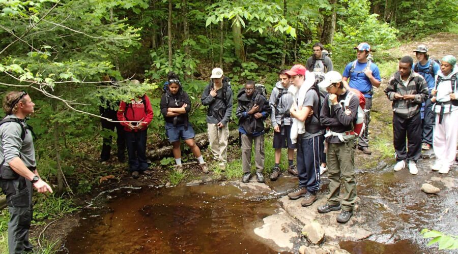 A group of people in outdoor clothing listens to a guide by a stream in a forested area. A group of people in outdoor clothing listens to a guide by a stream in a forested area.