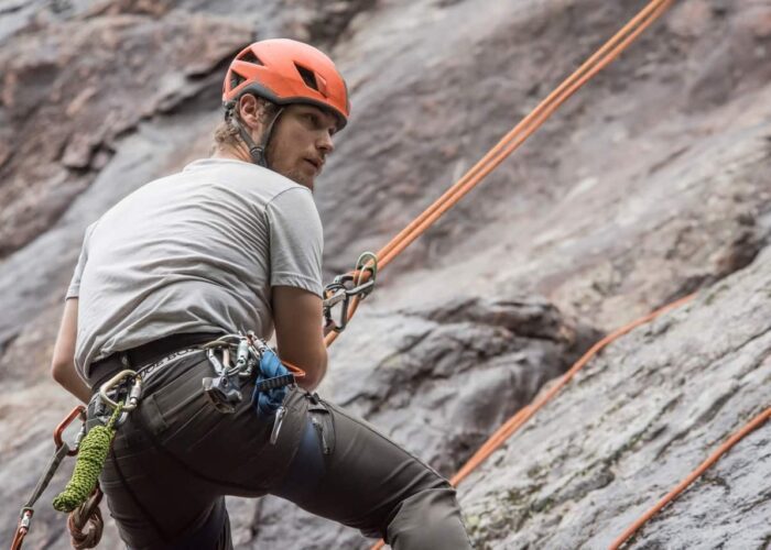 A person wearing a helmet and climbing gear is rock climbing outdoors, secured with ropes against a rocky surface.