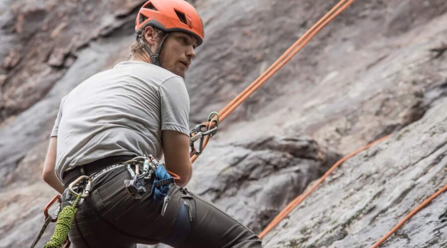 A person wearing a helmet and climbing gear is rock climbing outdoors, secured with ropes against a rocky surface.