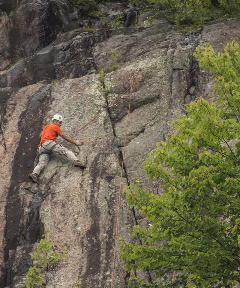 Une personne portant un casque et une chemise orange escalade une paroi rocheuse, profitant d'une aventure d'escalade au milieu des arbres verdoyants de Mont-Tremblant.