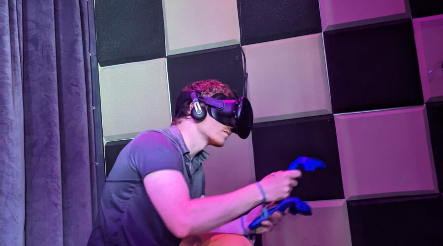 A person wearing a virtual reality headset and headphones holds two blue controllers in front of a soundproofed wall. A person wearing a virtual reality headset and headphones holds two blue controllers in front of a soundproofed wall.