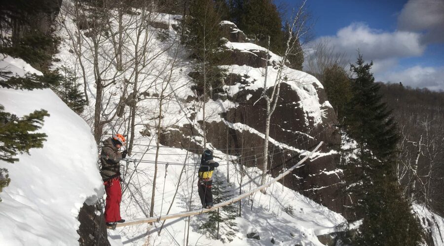 Two people in winter gear cross a narrow rope bridge over a snowy, rocky cliffside surrounded by trees. Two people in winter gear cross a narrow rope bridge over a snowy, rocky cliffside surrounded by trees.