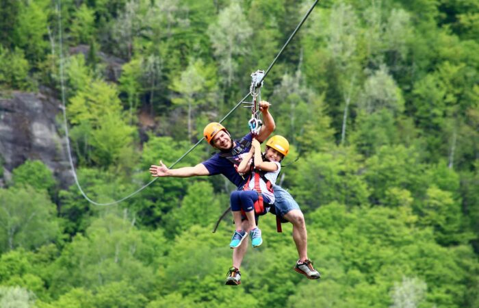 Two people wearing helmets and harnesses ride together on a zipline above a forested area, with trees and rocky terrain in the background.