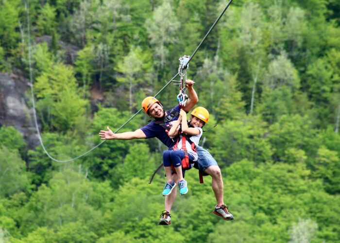 Two people wearing helmets and harnesses ride together on a zipline above a forested area, with trees and rocky terrain in the background.