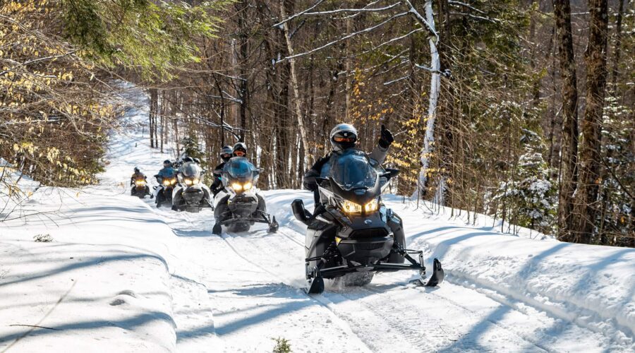 A group of people ride snowmobiles through a snowy forest trail, surrounded by tall trees with sparse snow on the branches.