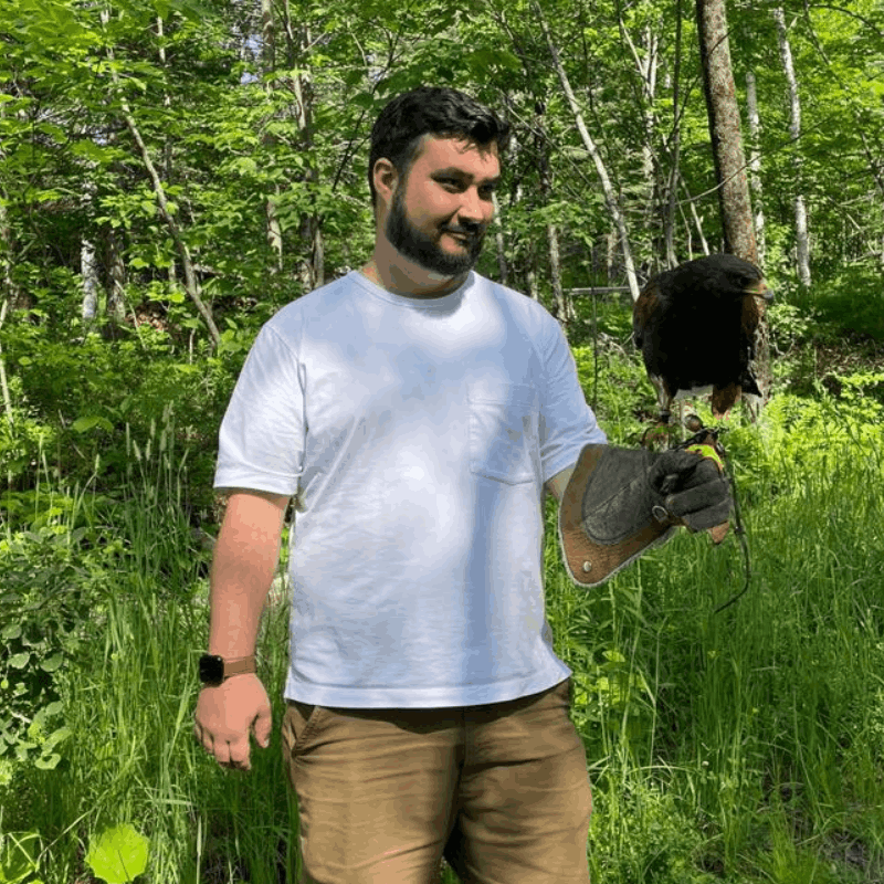A man wearing a white shirt and brown pants holds a bird of prey on a gloved hand in a green, wooded area. A man wearing a white shirt and brown pants holds a bird of prey on a gloved hand in a green, wooded area.