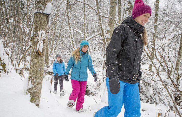Trois personnes portant des vêtements d'hiver se promènent avec le sourire dans une forêt enneigée de Tremblant, profitant d'une chute de neige fraîche - parfaite pour une journée de location de raquettes.