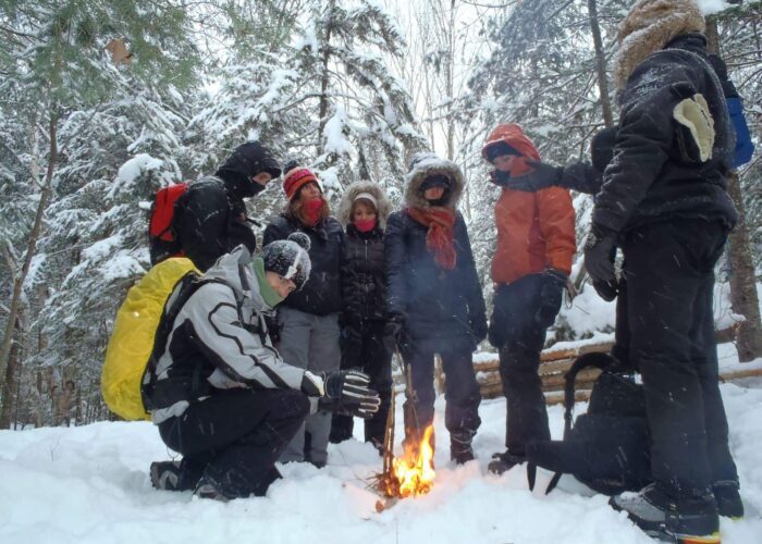 A group of people dressed in winter clothing stand around a small campfire in a snowy forest.