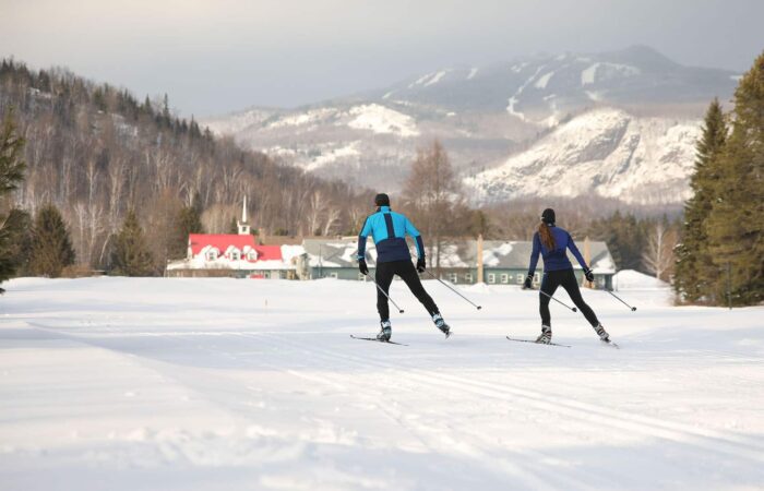 Two people cross-country skiing on a snowy trail with buildings and snow-covered mountains visible in the background.
