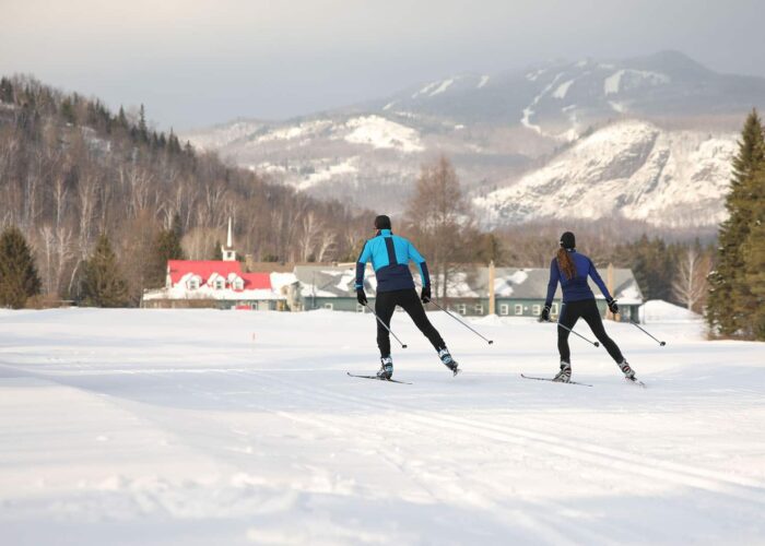 Two people cross-country skiing on a snowy trail with buildings and snow-covered mountains visible in the background.