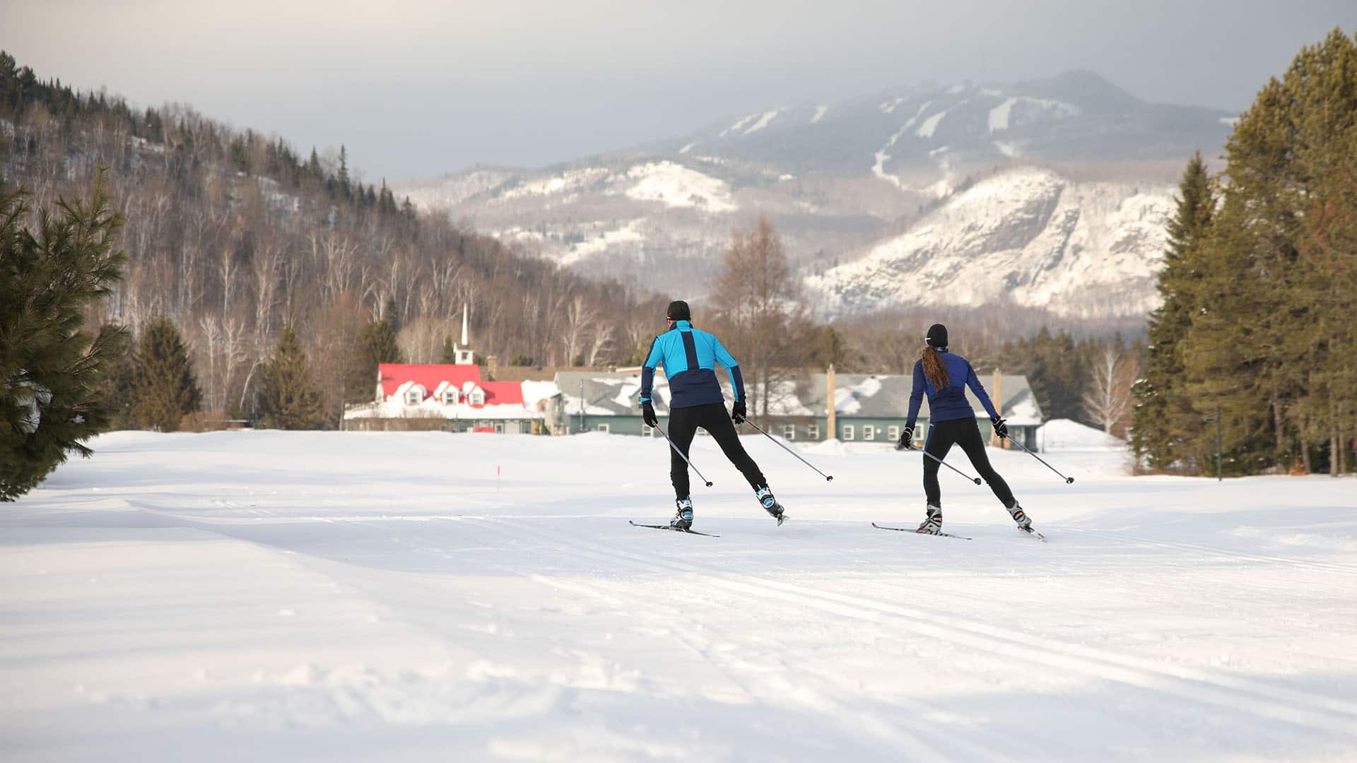 Deux personnes faisant du ski de fond sur une piste enneigée avec des arbres, des bâtiments et des montagnes en arrière-plan, profitant de la location de ski de fond et de l'accès Domaine Saint-Bernard.