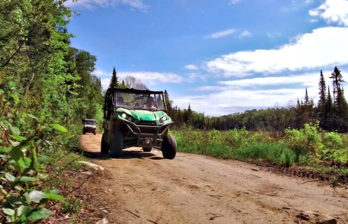Un véhicule tout-terrain vert roule sur un sentier de terre entouré d'arbres et de verdure sous un ciel partiellement nuageux - une aventure idéale pour un week-end entre filles au Mont-Tremblant.