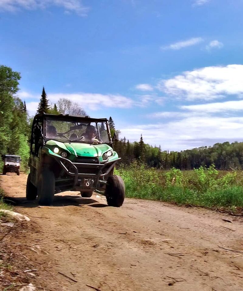 A green all-terrain vehicle drives on a dirt trail through a forested area under a partly cloudy sky.