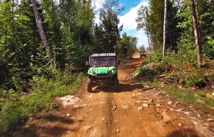 An off-road vehicle drives on a rugged dirt trail through a forested area under a blue sky, with another vehicle visible in the background.