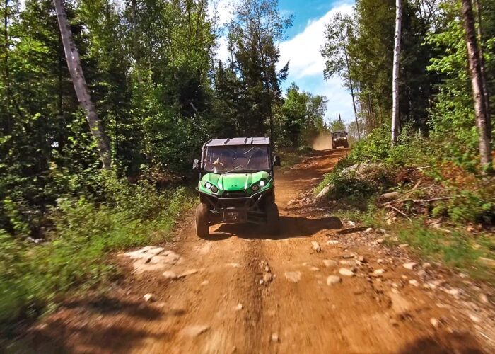 An off-road vehicle drives on a rugged dirt trail through a forested area under a blue sky, with another vehicle visible in the background.