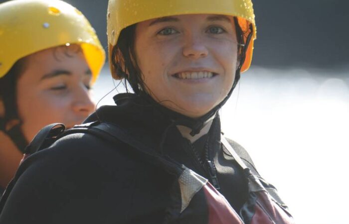 Two people wearing yellow helmets and life jackets stand outdoors near water, with one person smiling at the camera.