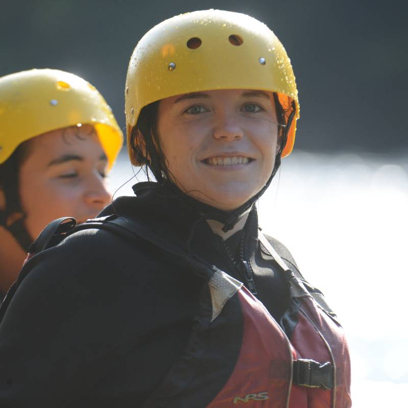 Two people wearing yellow helmets and life jackets stand outdoors near water, with one person smiling at the camera. Two people wearing yellow helmets and life jackets stand outdoors near water, with one person smiling at the camera.