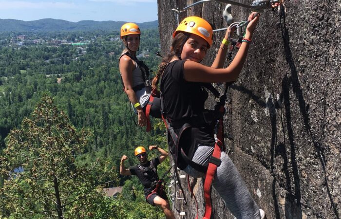 Three people wearing helmets and harnesses climb a vertical rock face, with a forested landscape and hills visible in the background.