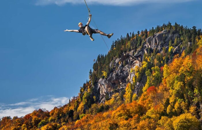 A person ziplining in front of a forested, rocky hillside with vibrant autumn foliage under a clear blue sky. A person ziplining in front of a forested, rocky hillside with vibrant autumn foliage under a clear blue sky.