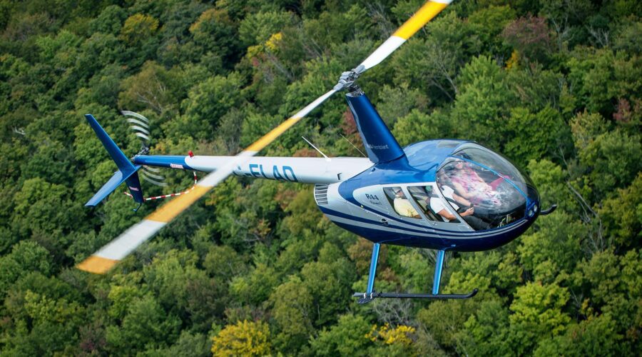 A blue and white helicopter flies over a dense, green forest, with its rotor blades in motion and two people visible inside the cockpit.