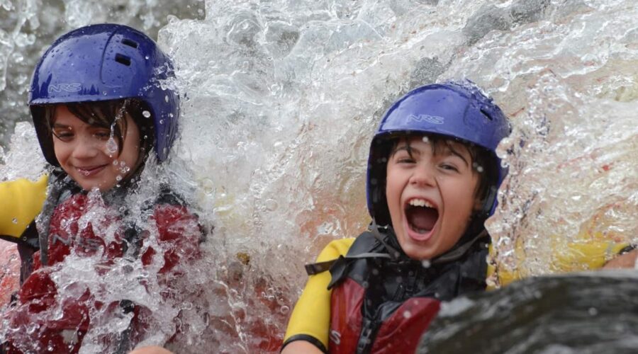 Two children wearing helmets and life jackets are splashed with water while whitewater rafting; one smiles and the other shouts with excitement.