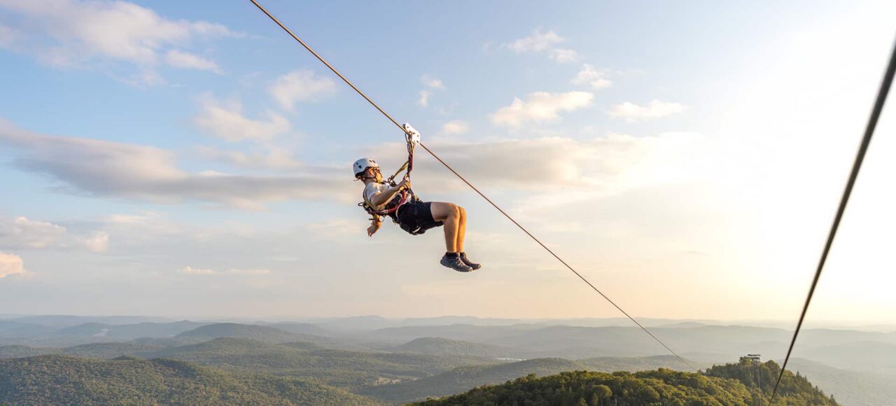 Person ziplining above a forested mountain landscape under a blue sky with scattered clouds, with distant hills visible in the background.
