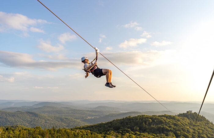 Person ziplining above a forested mountain landscape under a blue sky with scattered clouds, with distant hills visible in the background. Person ziplining above a forested mountain landscape under a blue sky with scattered clouds, with distant hills visible in the background.