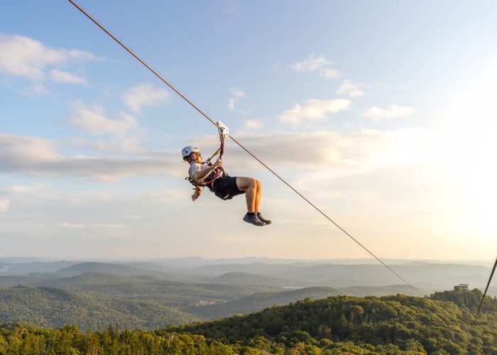 Person ziplining above a forested mountain landscape under a blue sky with scattered clouds, with distant hills visible in the background.