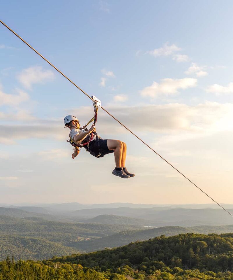 Person ziplining above a forested mountain landscape under a blue sky with scattered clouds, with distant hills visible in the background.
