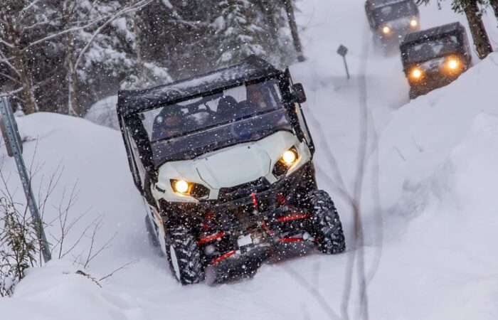 Four off-road vehicles drive through a snowy forest trail during snowfall, with headlights on and snow covering the ground and trees.