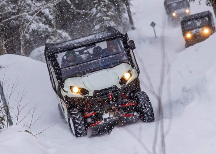 Four off-road vehicles drive through a snowy forest trail during snowfall, with headlights on and snow covering the ground and trees.
