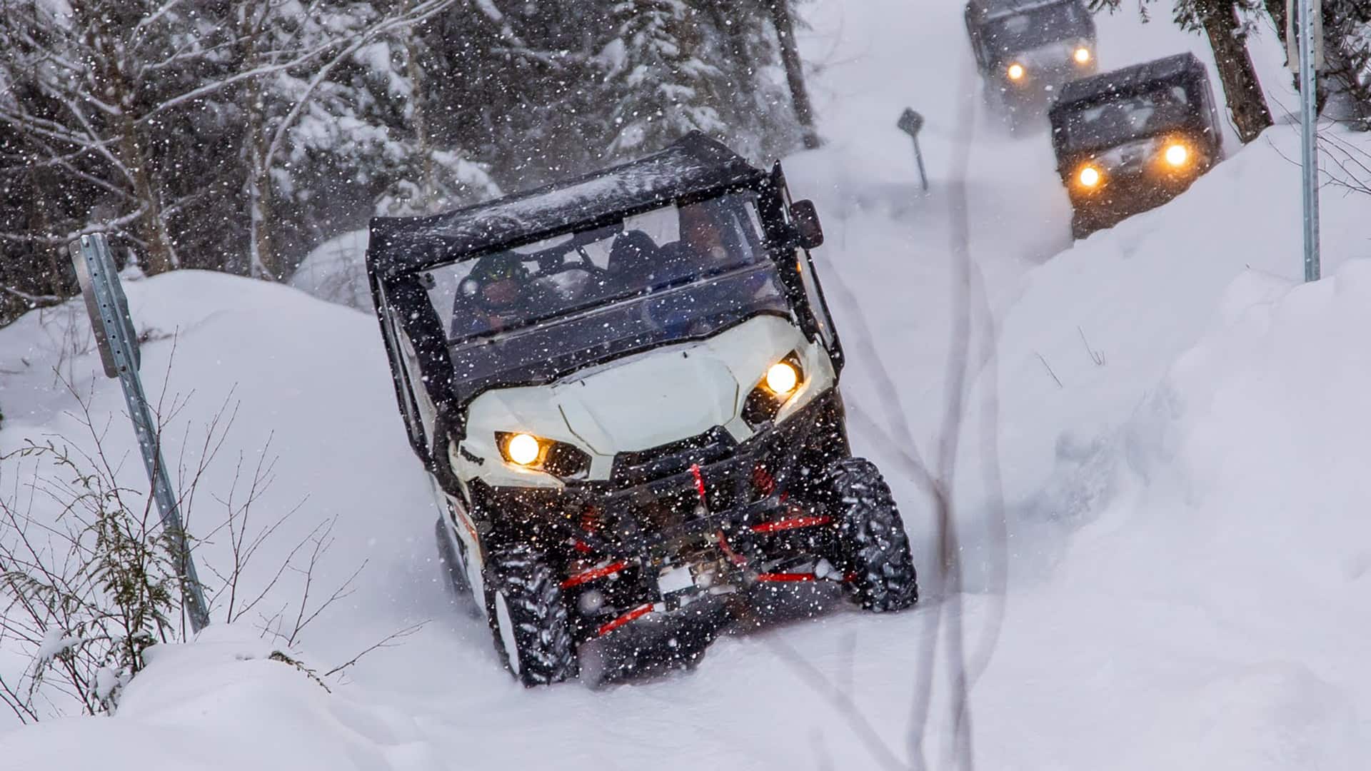 Four off-road vehicles drive through a snowy forest trail during snowfall, with headlights on and snow covering the ground and trees.